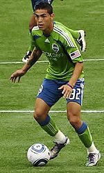 Two men in blue and green soccer uniforms. One has the ball at his feet and is looking upfield.