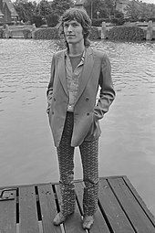 A long-haired man standing on a platform by a body of water